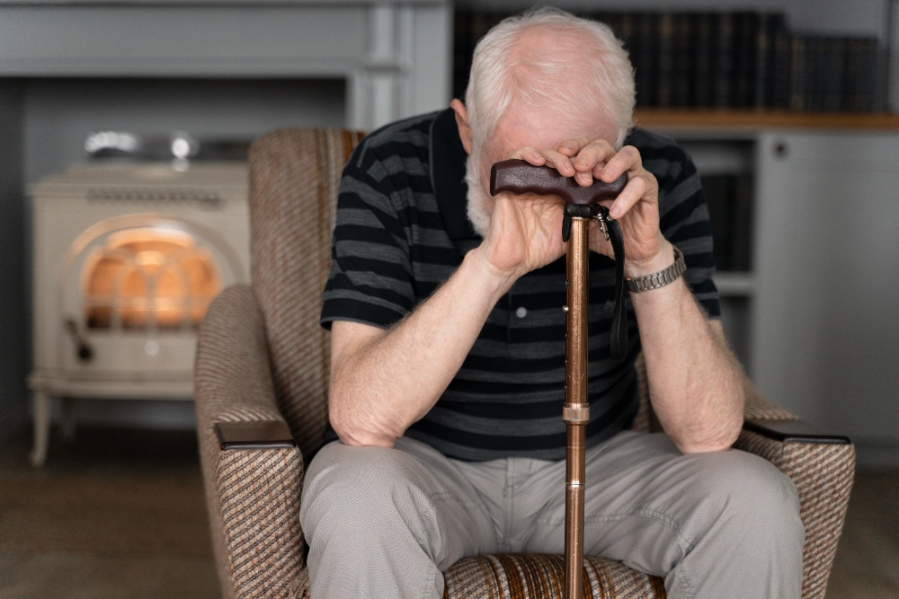 Elderly man rests forehead on cane handle, seated by fireplace.